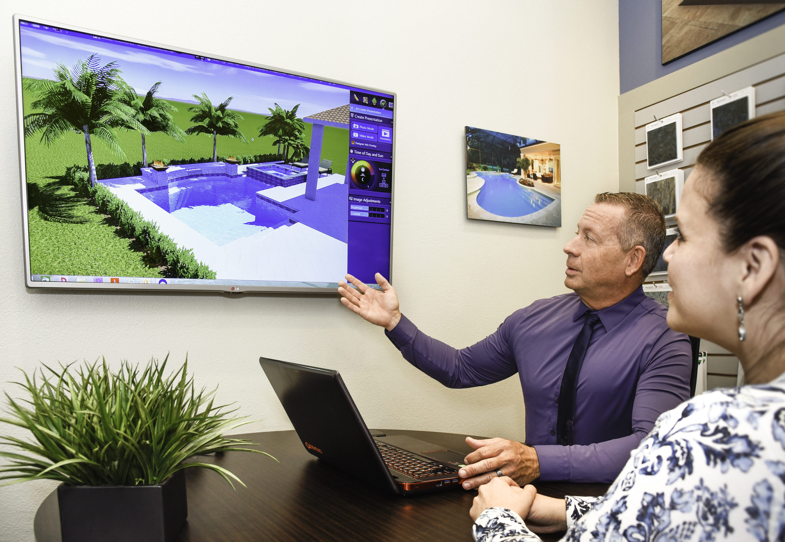 a man in a purple shirt and tie sitting at a table with a laptop and a plant in front of him